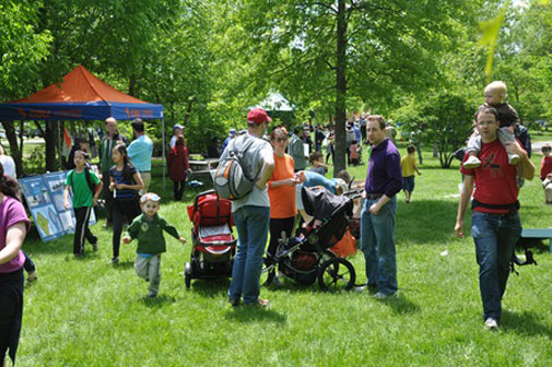 A grassy field with trees, in it are families looking at the info tables of Fresh Pond Day