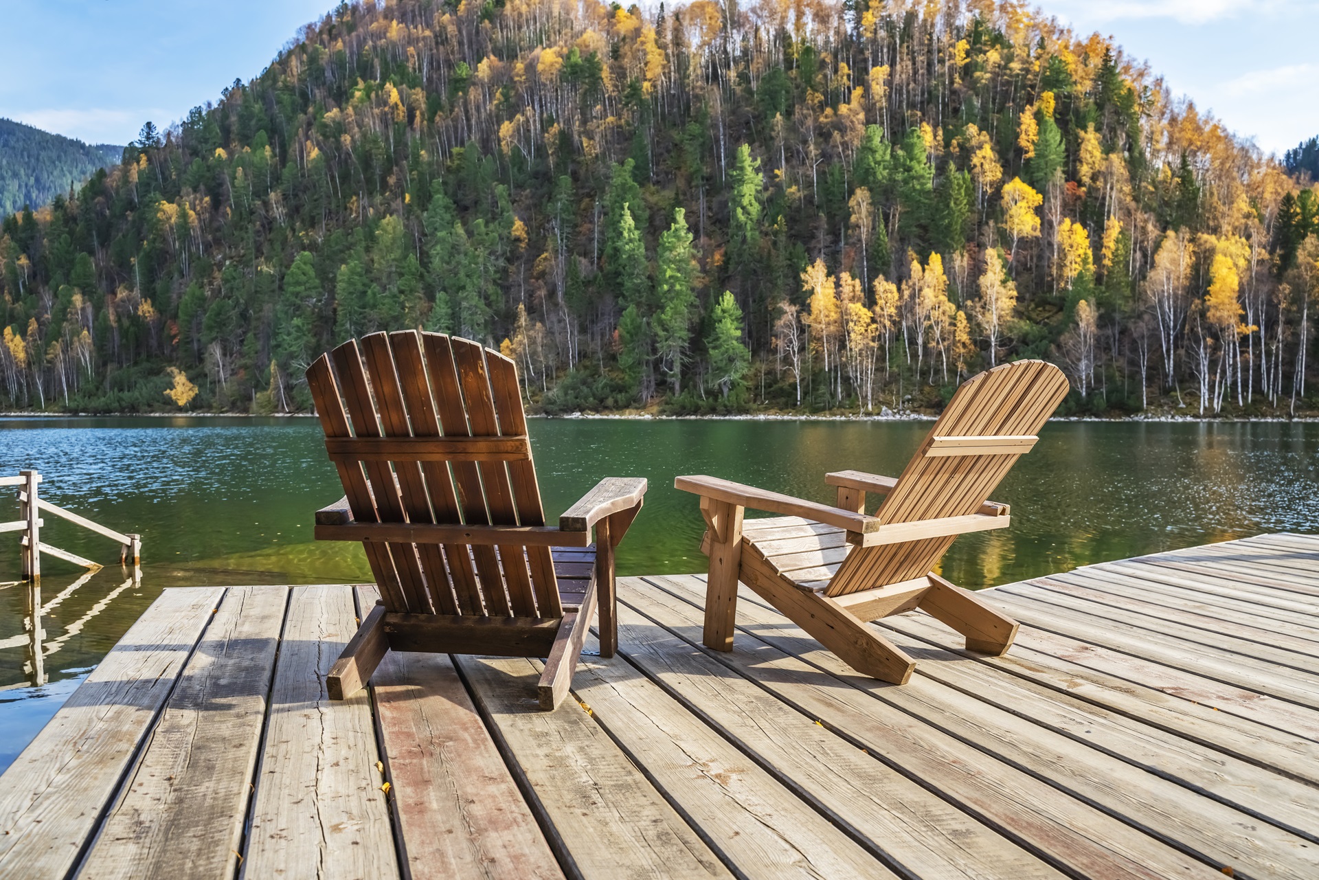 A dock on a lake with two Adirondack chairs facing out on the water, a mountain in the distance with green and yellow trees