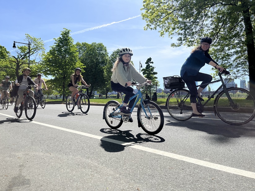 A family rides their bikes down Memorial Drive through the middle of the street. The Boston skyline is in the background.