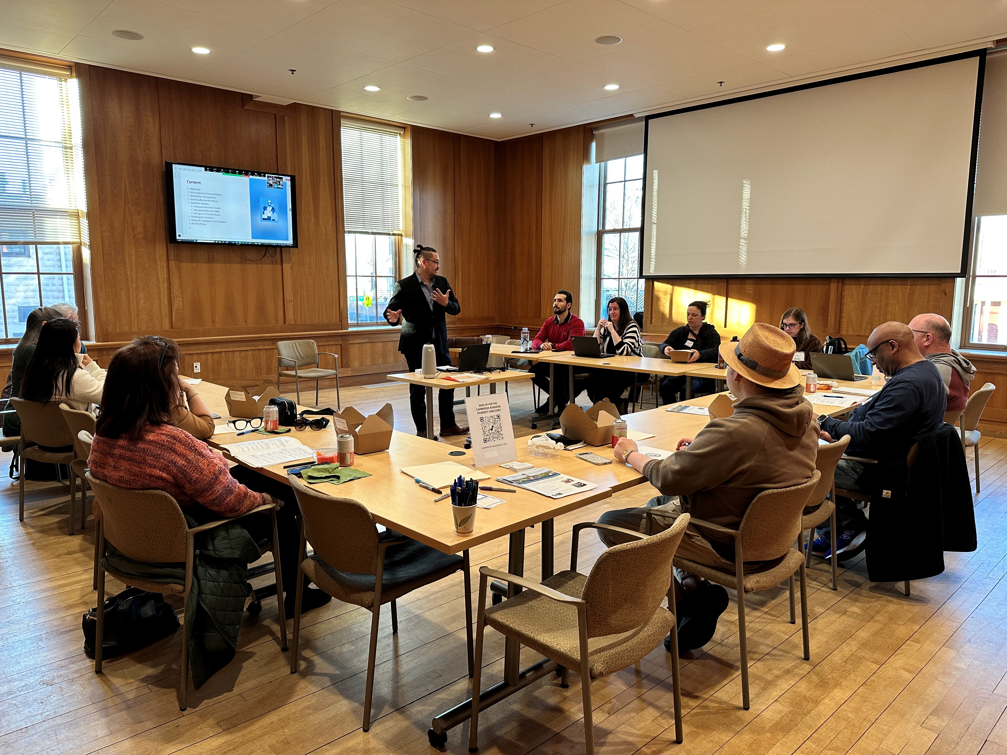 workshop participants sit around a table looking up at a presenter