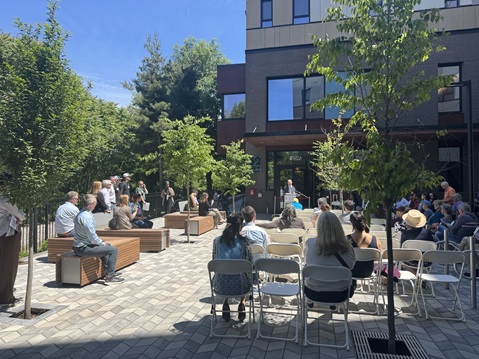 A crowd of people seated outside on a patio with seedlings and sunshine, watching a speaker at a podium at 52 New Street