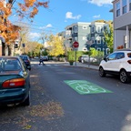 Photograph of a one-way residential street with a lime green rectangle and white bike icon painted in the middle. A postal worker is using a crosswalk at a raised intersection in the background.