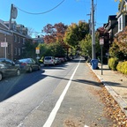 A photograph of a one-way residential street with houses and parked cars at left and a sidewalk and marked bike lane on the right