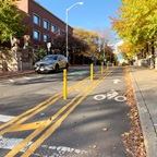 A photograph of a one way street coming down a hill with parked cars on the far left, a vehicle lane with a lime green bicycle icon in the center, yellow flexible dividers on the right, and a separated bike lane on the far right headed in the opposite direction