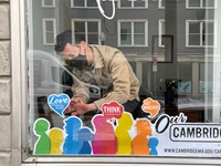 Person with brown hair wearing mask putting up colorful window decal in storefront.