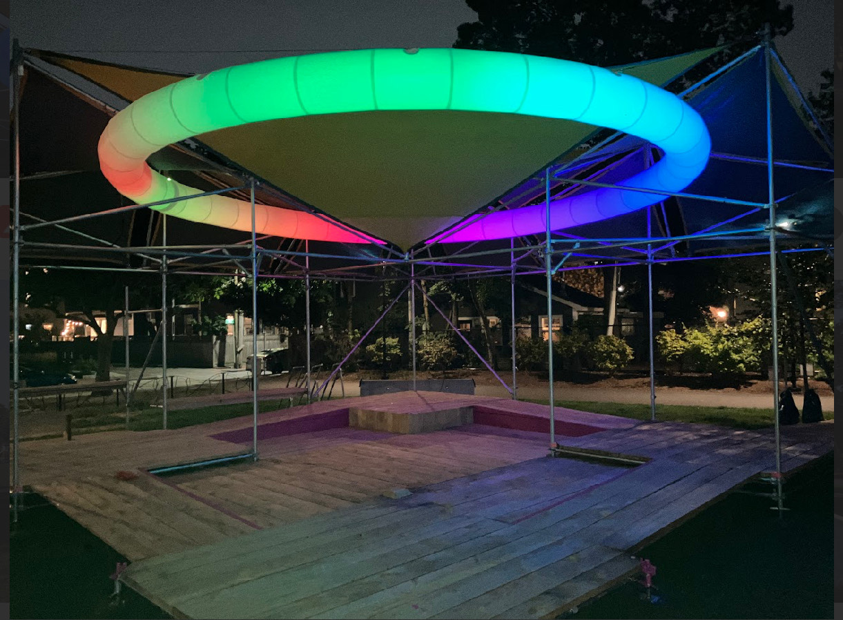 Picture of SHADE installation in Sennott Park at night. Structure features a large lit and glowing circle on the ceiling with a rainbow of bright colors.