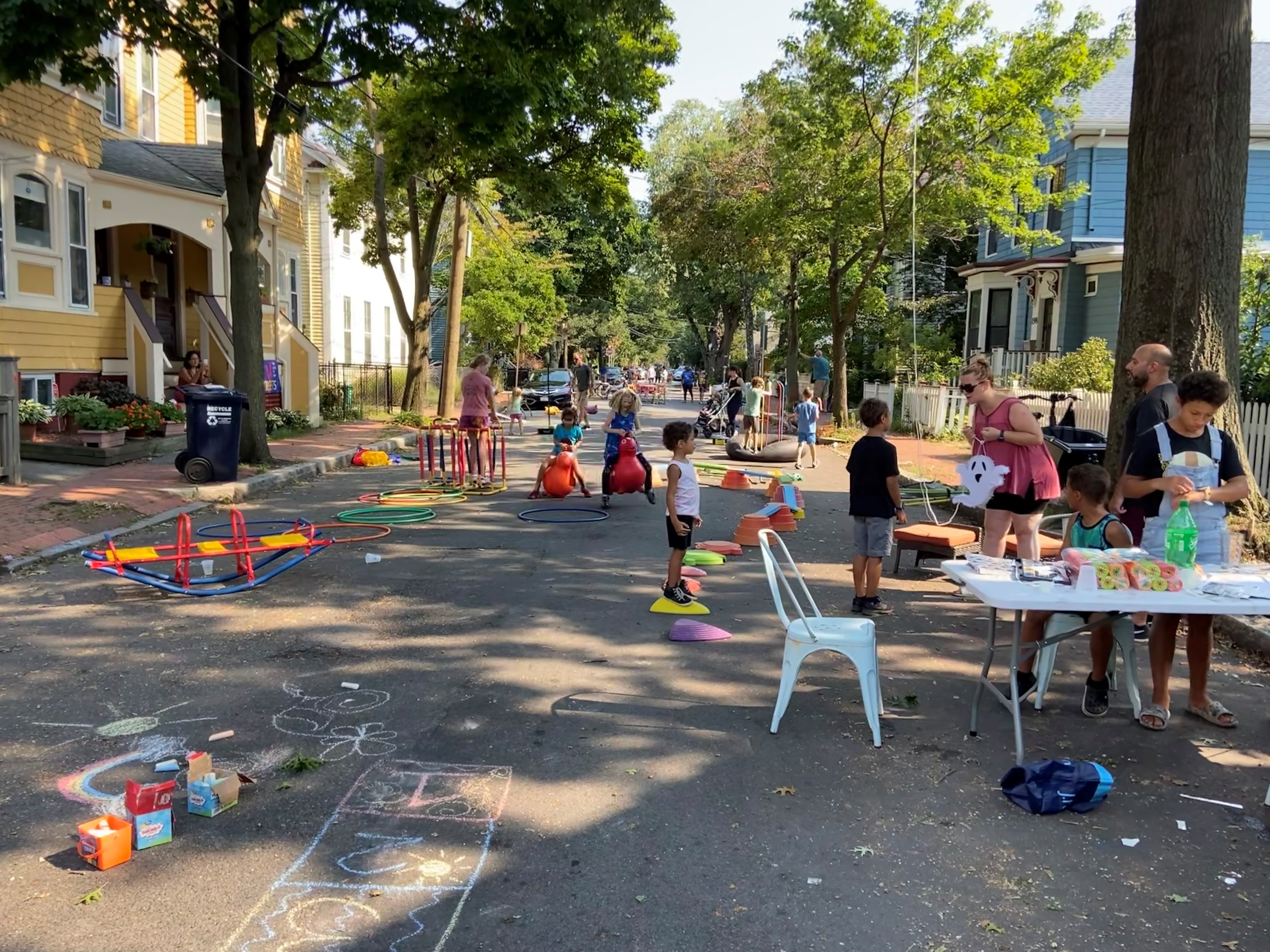 A photo of young people playing on a city street. In the foreground one young person is playing with colorful blocks on a folding table.