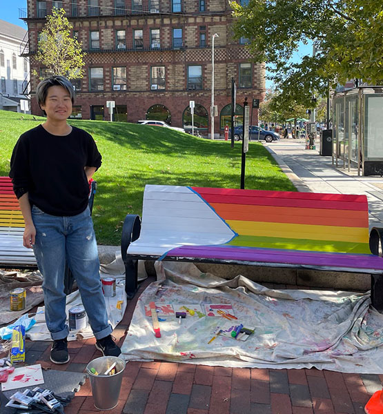 Person standing next to a bench that has been freshly painted with the rainbow flag