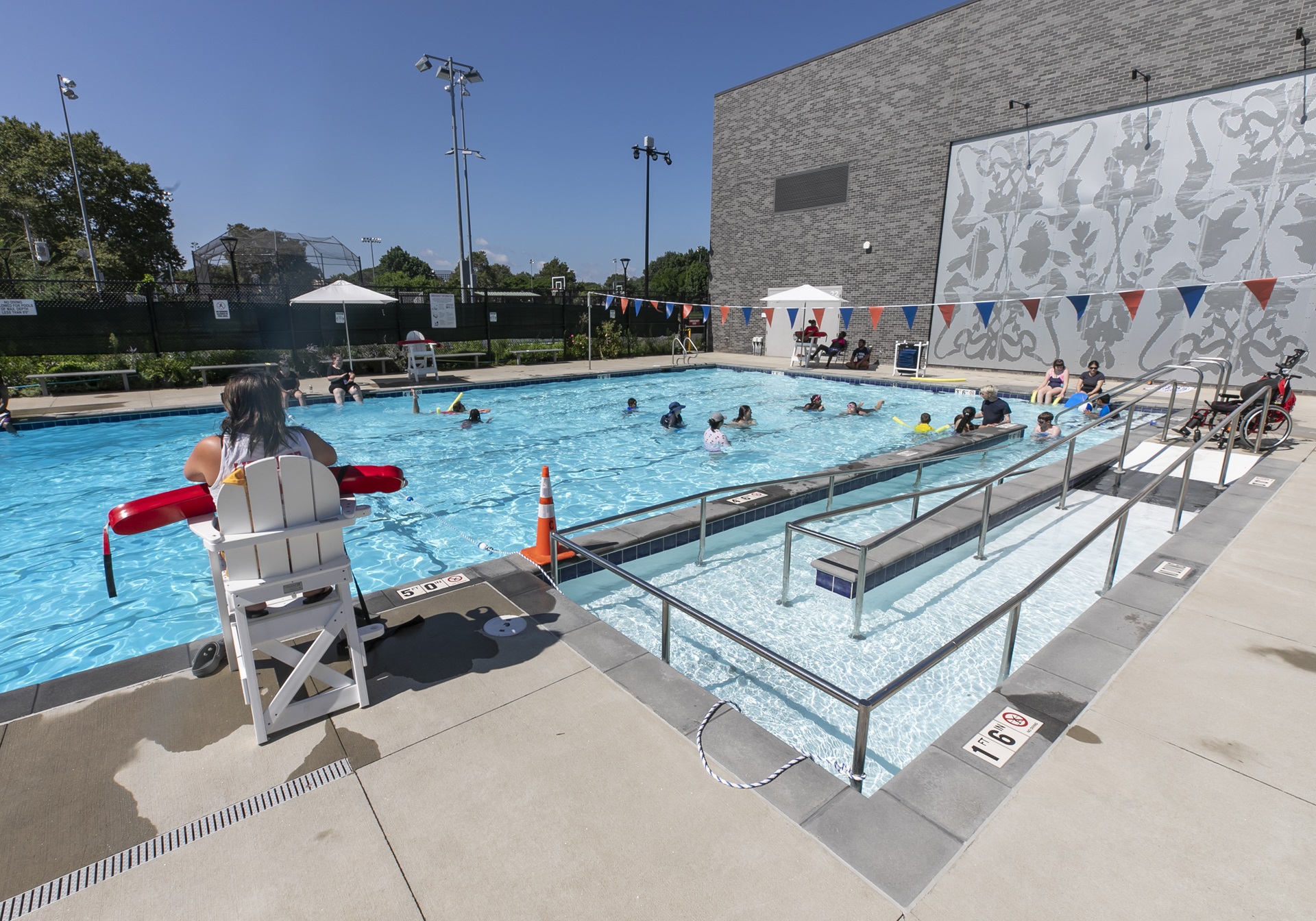 Gold Star Pool's lap pool is ADA compliant. Patrons swim in the shallow end and a wheelchair is parked at the top of the wheelchair ramp. A lifeguard oversees the swimmers.