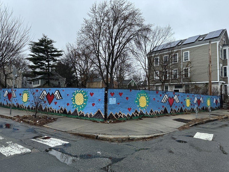 A lot at 35 Cherry Street where affordable homeownership units will be built. A fence surrounds it that's decorated with a blue background and designs of mountains, hearts with wings, and suns.