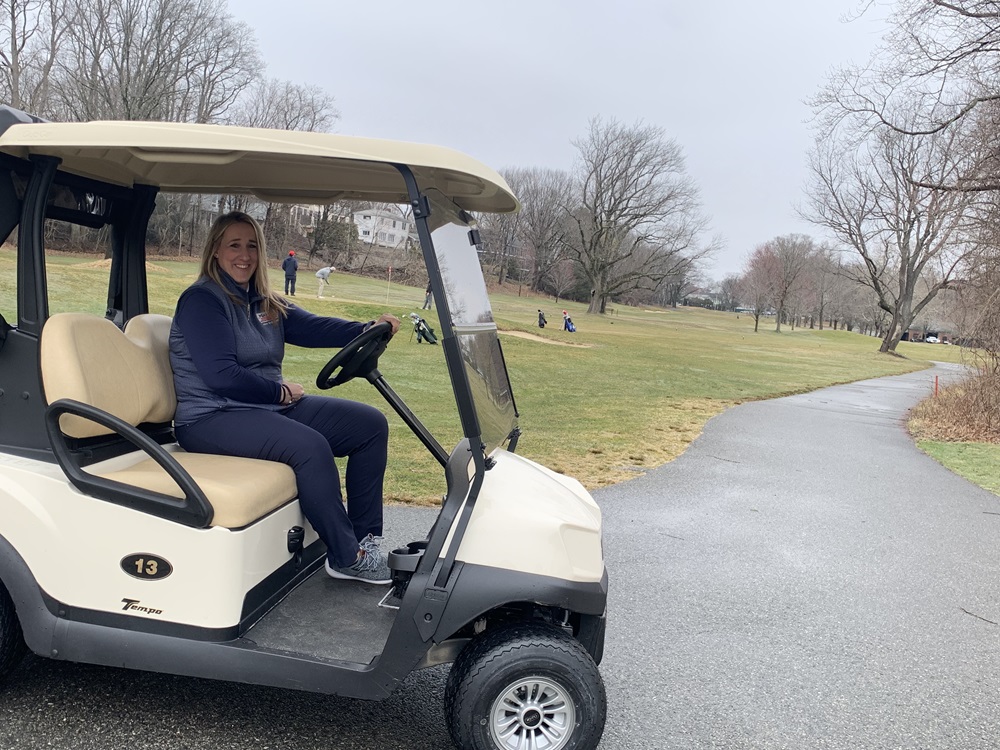 Golf Director Jo-Anna Krupa on a golf cart with golfers on the green behind her.