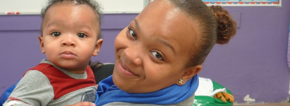 A mother smiling at the camera while holding an infant child in a classroom setting.
