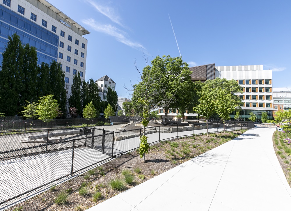A wide walkway passes through a landscaped urban area with trees, plants, benches, and surrounding modern office buildings under a clear blue sky.”