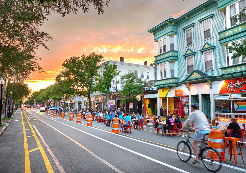 Landscape photo of Inman Square dining establishments, a resident riding alongside the road on a bike with a glowing sunset in the background.