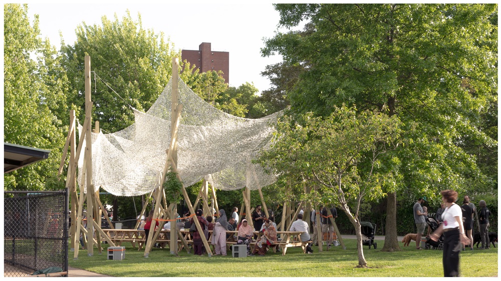 Cambridge residents sit at picnic tables in a local green space, covered by a shade structure.