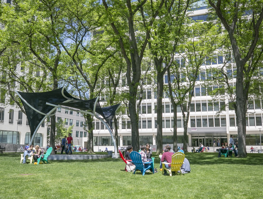 Cambridge residents enjoy sitting in chairs under a shade structure in Kendall Square.