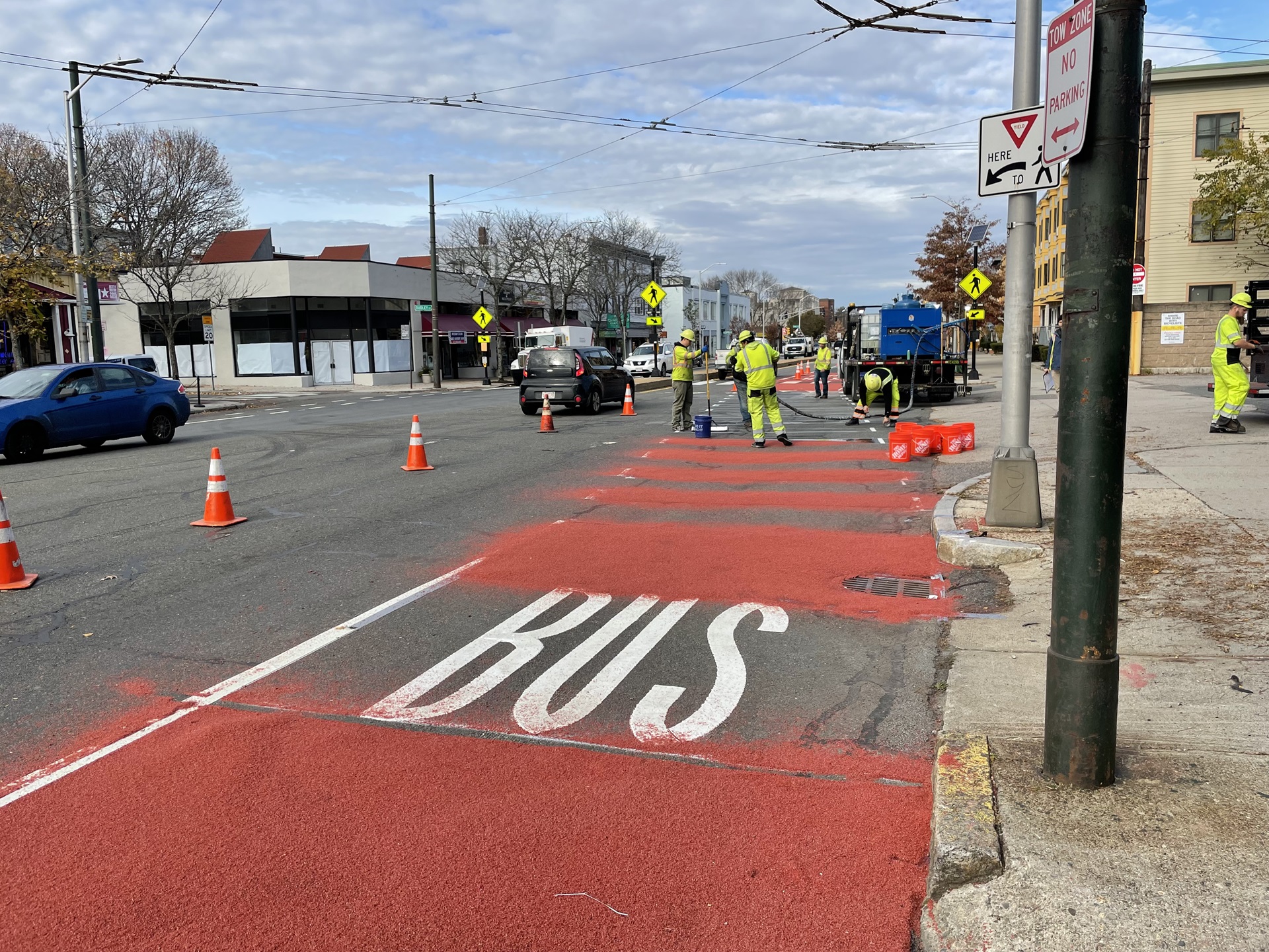 A crew of five people in neon yellow installs a bus lane by painting the lane red. To the left, cones temporarily separated the crew and lane from the rest of the street.
