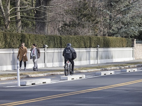 A cyclist rides in the two-way separated bike lane on Brattle Street. On the sidewalk next to the cyclist, two women walk.