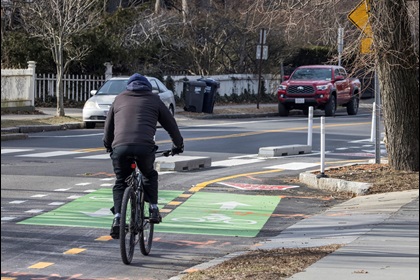 A photo from the Phase 1 project area shows a person on a bike traveling in the two-way bike lane, separated from the vehicle lane with concrete curbs. On the opposite side of the street, two cars are parked.