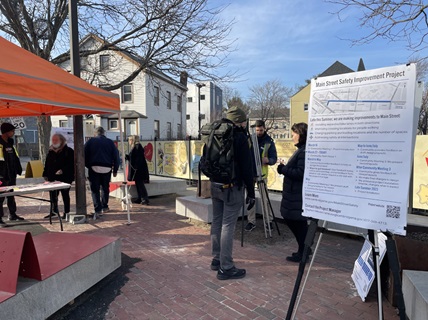 Seven people stand in a plaza of Main Street. Some residents are chatting with city staff, others are looking at posters. A poster in the foreground shows the timeline for the project, from March 2023 to late summer 2023.