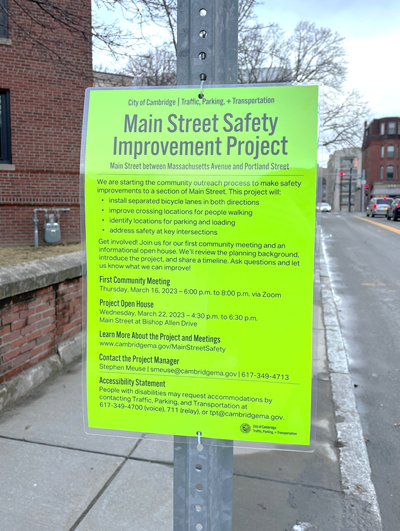 A bright yellow poster on a signpost announces the Main Street Project, first community meeting, and project open house.