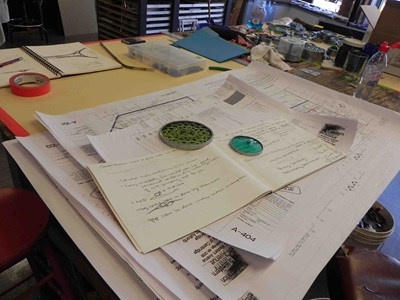 Photograph taken from a pre-fabrication review at an artist's studio showing notes, sketches, construction drawings, and sample materials.