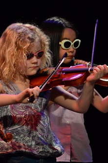 Young violinists perform together on stage wearing colorful sunglasses during Cambridge Open Studios.