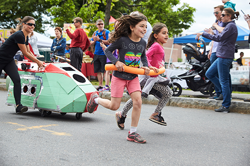 Two children race down a street pulling a green, wheeled sculpture that looks vaguely like a spaceship. The sculpture is also being pushed from behind by an adult. They race down an outdoors street.