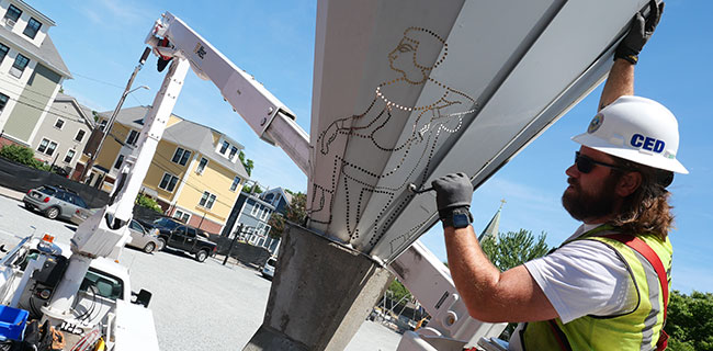 Michael Corcoran of the Cambridge Electrical Department removes a shade from John Tagiuri's "Endless Lamps" at Sennott Park, to help Cambridge Arts refurbish the public artwork so that it looks fresh when renovation of the park is completed, June 2022.