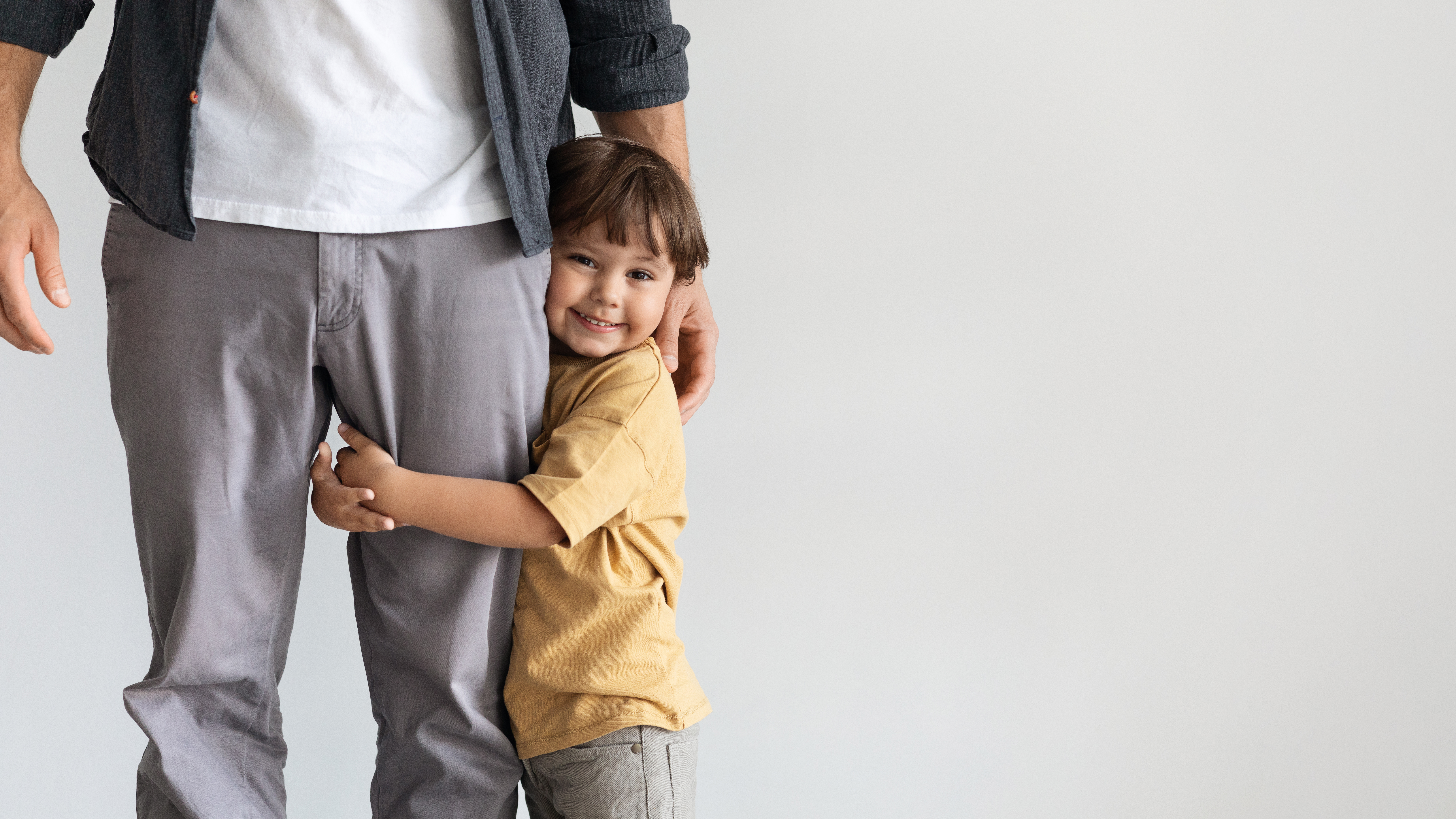 A toddler smiles at the camera and clings to their parent's leg