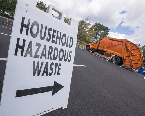 Photo of sign that reads "Household Hazardous Waste" with a directional arrow underneath; a bright orange DPW truck is visible in the background.