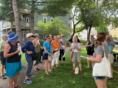 Photo of participants at the Mapping Feminist Cambridge: Inman Square Walking Tour; while standing in a group, the tour guide speaks to the group