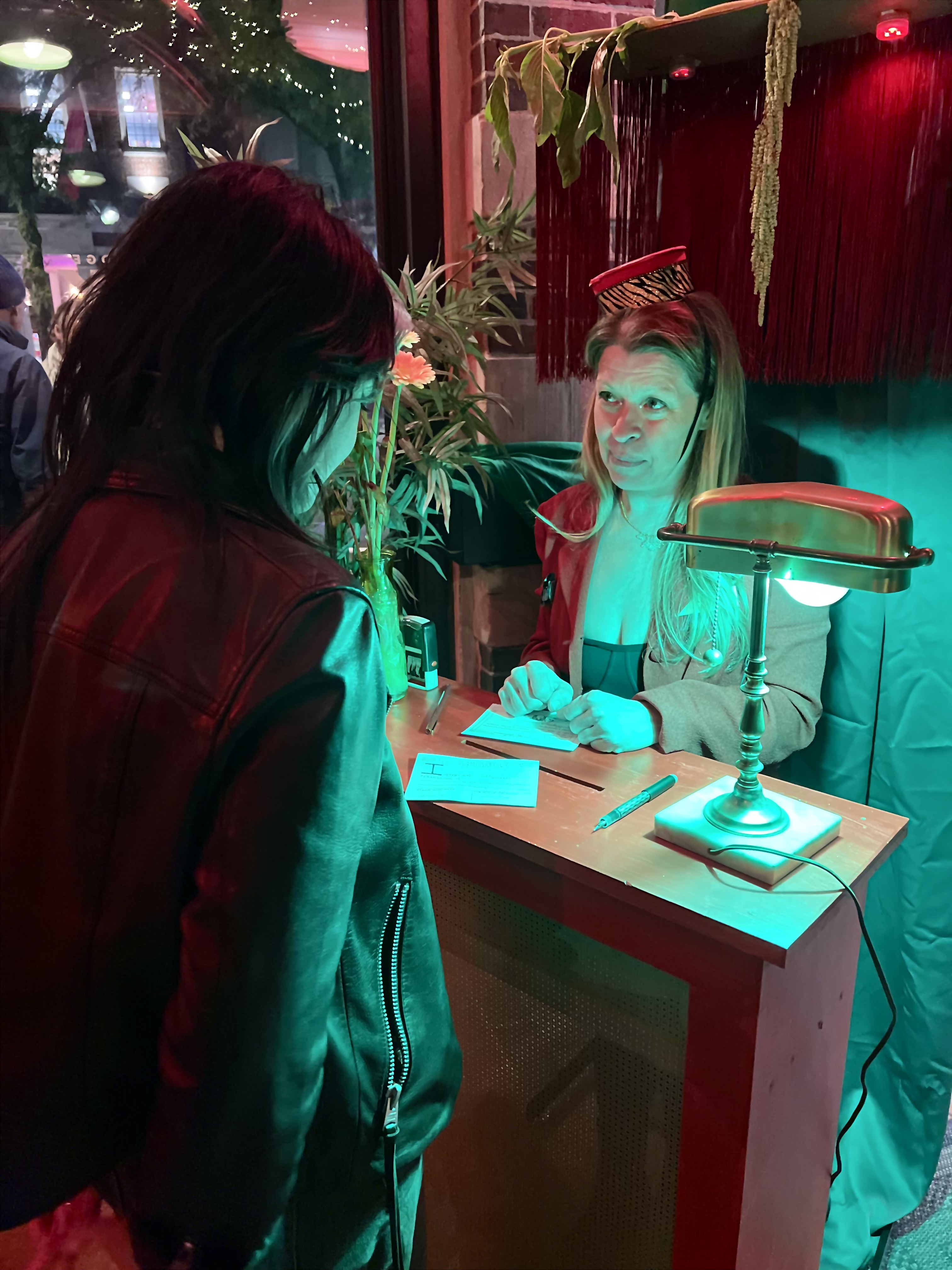 Person standing at a wooden counter in dim light providing information to another person who is visiting the Cambridge kiosk