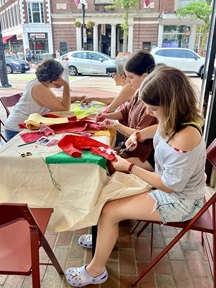Three people sitting at a table in the Cambridge Kiosk working with colorful pieces of fabric on a craft activity
