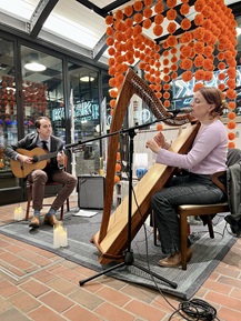 Person playing guitar and person playing the harp on a brick floor with tall glass windows and an art piece in the background consisting of long strings of hanging circles