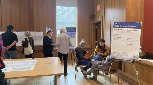 people siting indoors in a circle table with map on the left side, people standing in the background