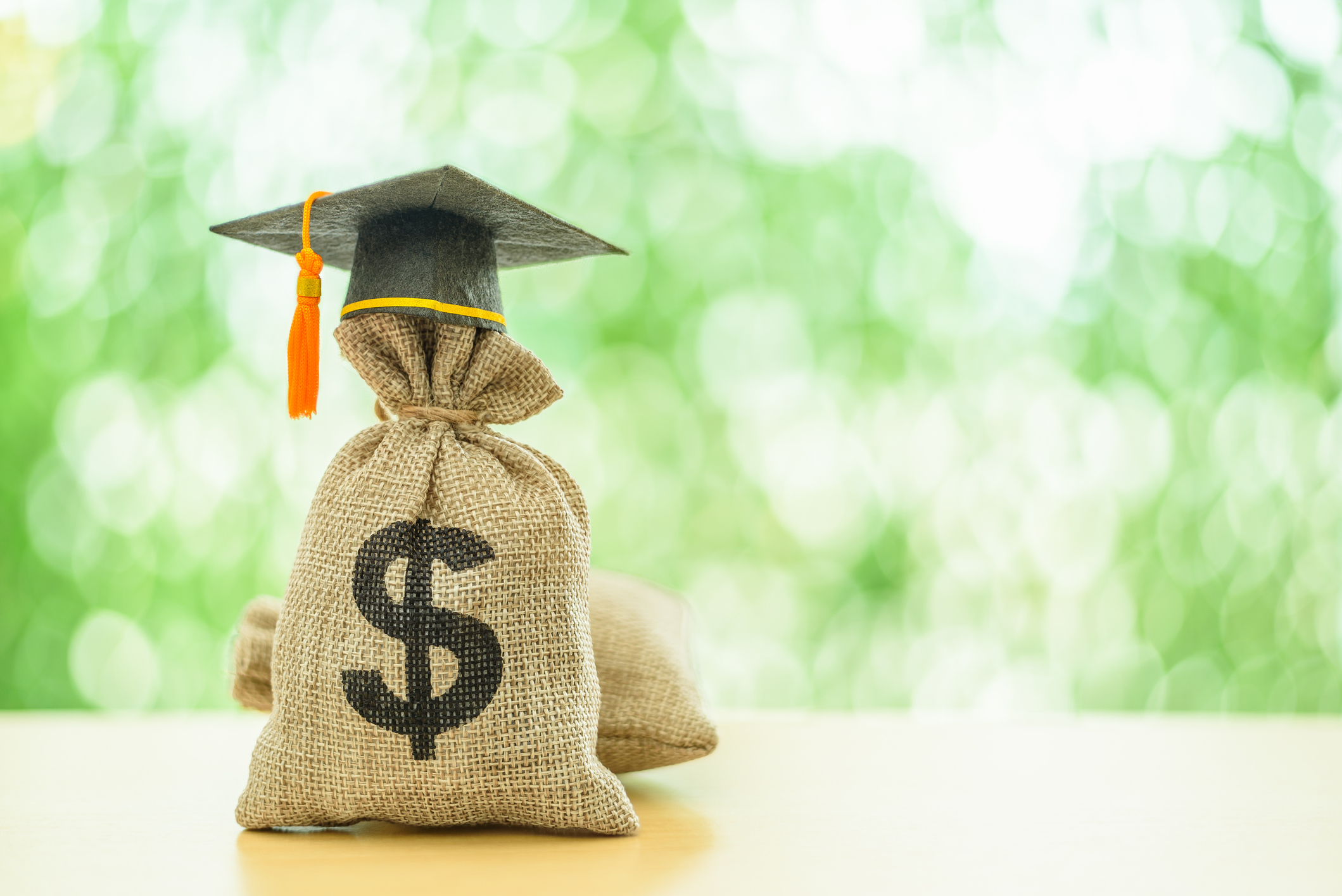 A burlap sack with a dollar sign written on it in black ink and wearing a black graduation cap sits on a wooden table with a natural green backdrop