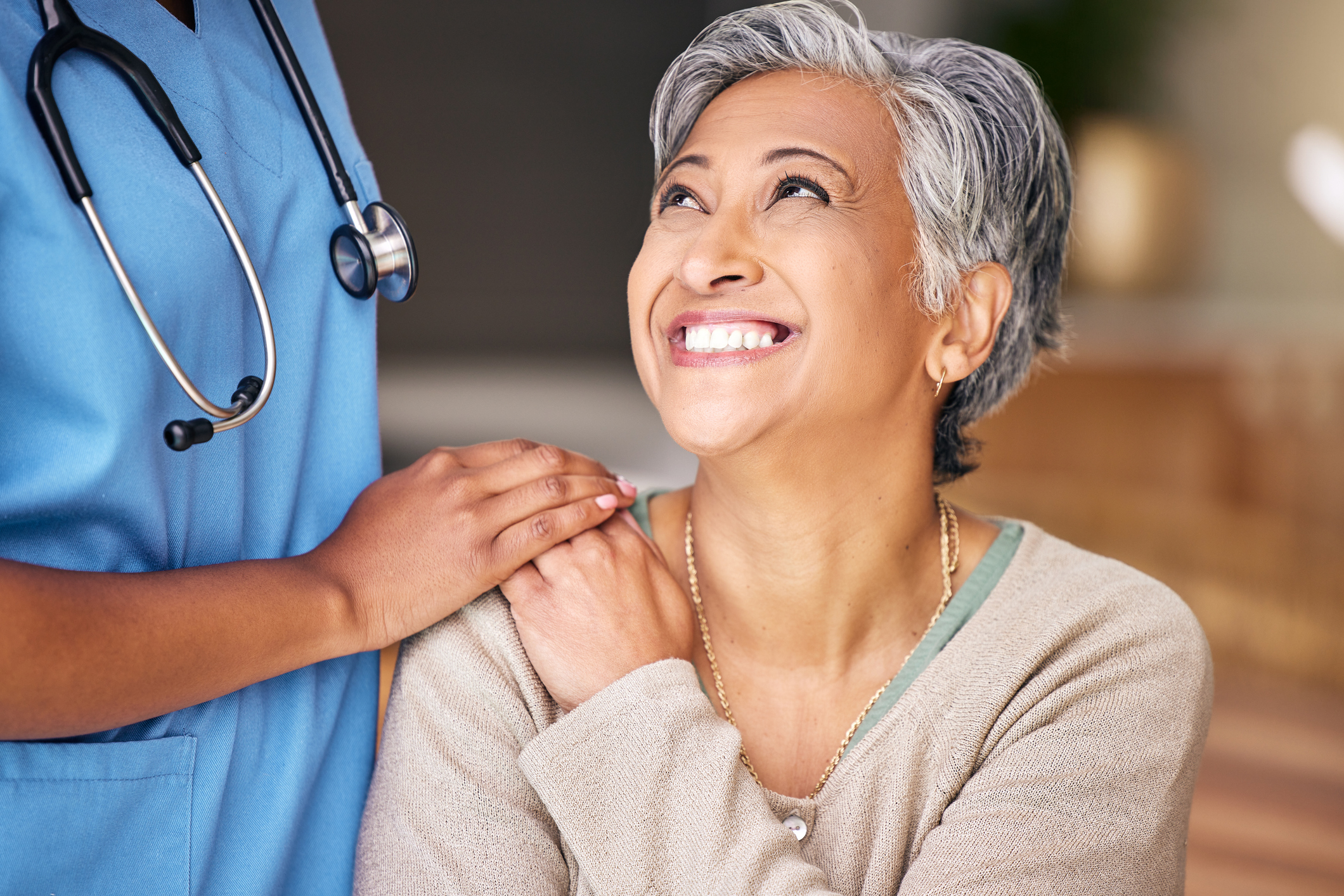 A woman looks up and smiles at a nurse or caregiver standing beside her with their hand on the woman's shoulder.