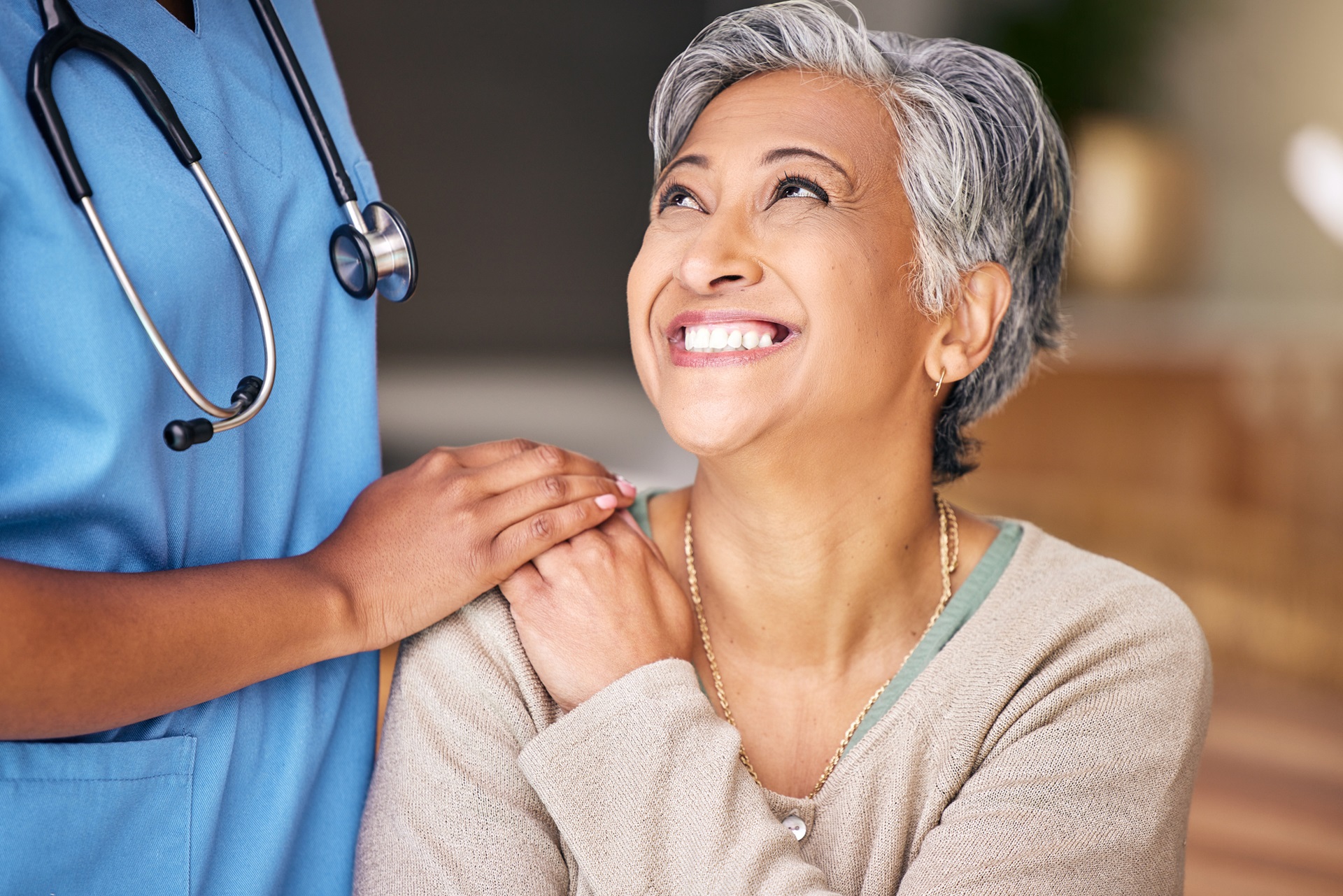 A woman looks up and smiles at a nurse or caregiver standing beside her with their hand on the woman's shoulder.