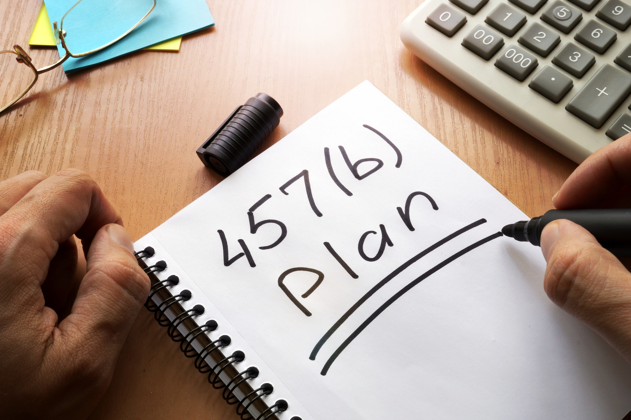 A cluttered office desk with a notebook on which a person has written and underlined '457(b) Plan' in black marker