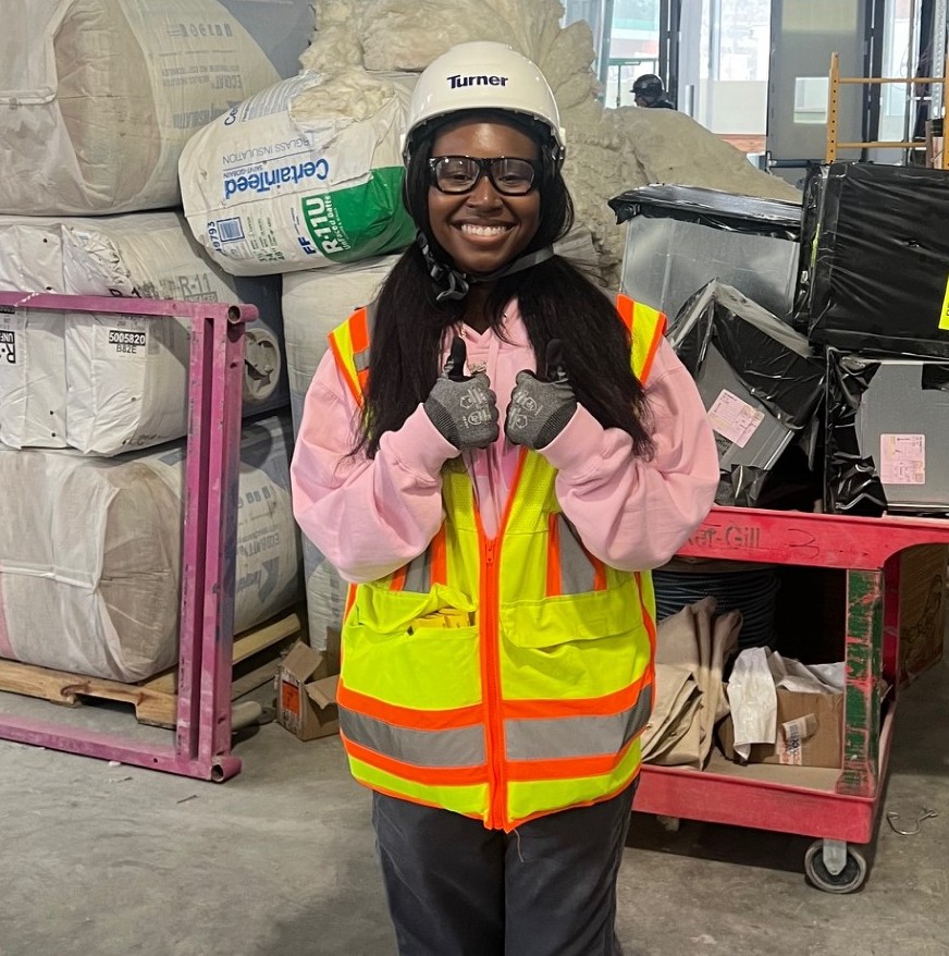 A young woman stands and smiles wearing a hard hat and construction vest