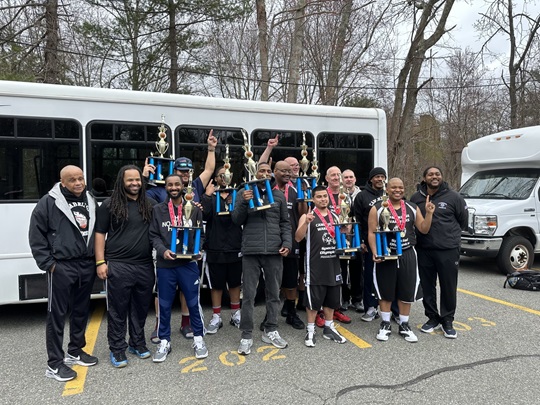 The Cambridge Warriors basketball team standing in front of a white bus, holding trophies and wearing medals. They are dressed in black basketball uniforms. They are celebrating a victory outdoors, with trees in the background.