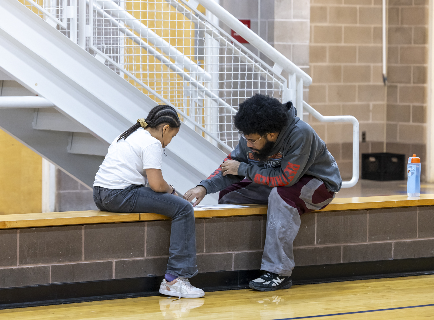 A Cambridge Youth Programs (CYP) staff member guides a young person through an activity. Both are seated on a bench in an indoor basketball court.