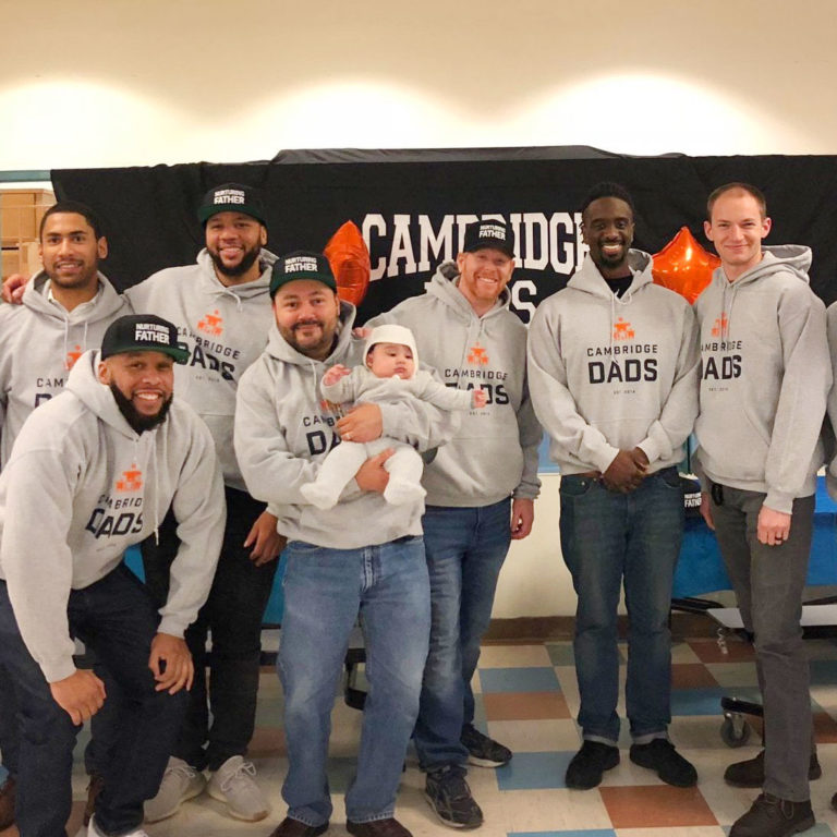 A group of men wearing Cambridge Dads sweatshirts pose for a photograph. One of the men is carrying an infant.