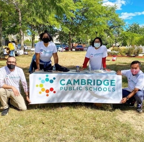 Four volunteers standing around a table in a park with a Cambridge Public Schools banner