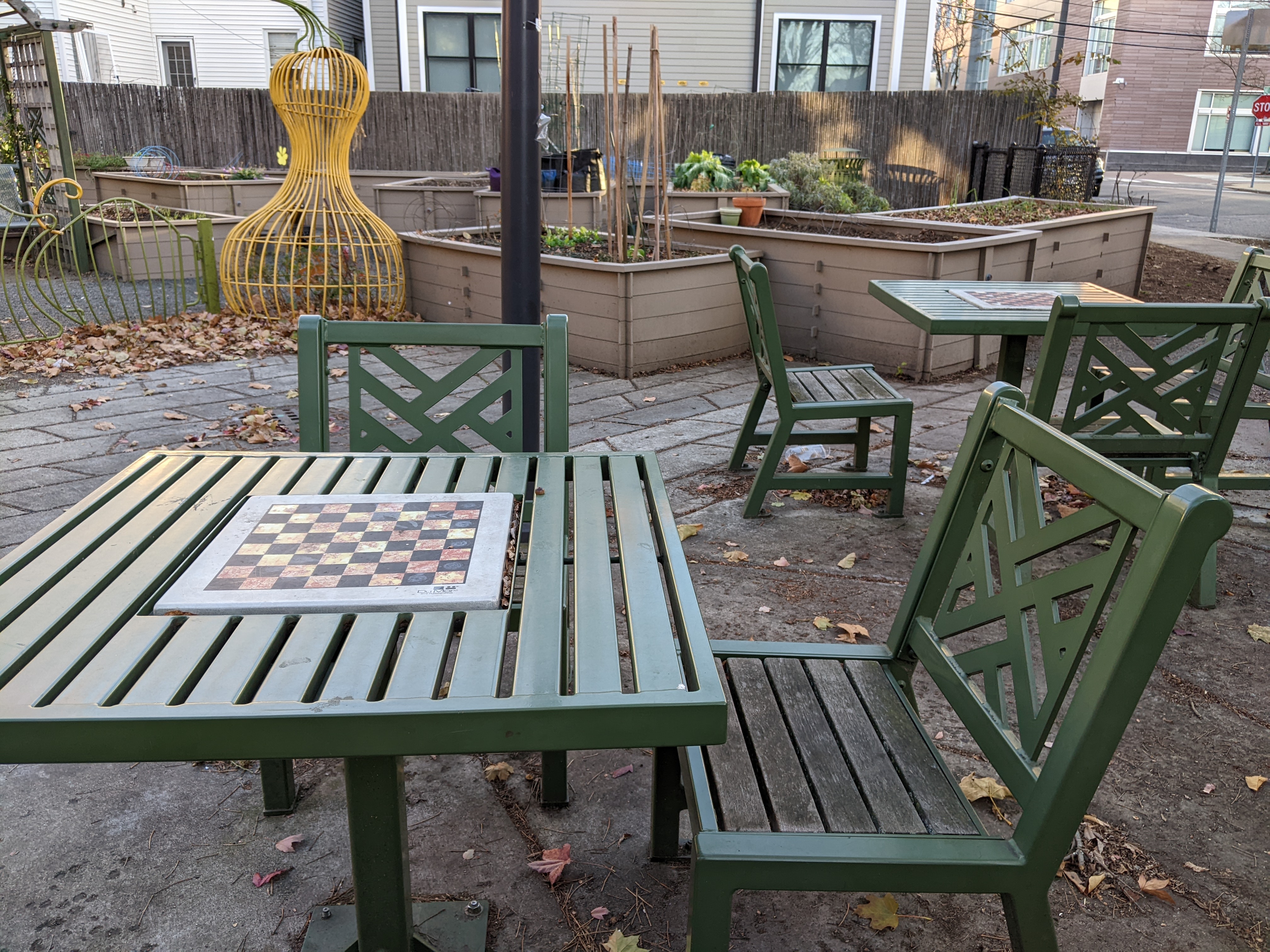 Two tables with embedded chess boards in a park next to a community garden.