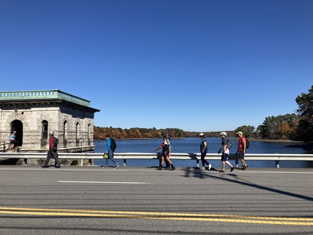 People walk along the side of a road before entering the Hobbs Brook Gatehouse, a white stone building. Blue water and blue sky are in the background.