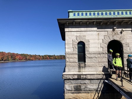 People enter and exit the Hobbs Brook Gatehouse, a white stone building that juts out into the reservoir. Blue water and colorful trees are seen to the left.
