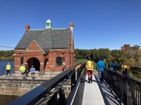 People walk across a metal bridge to the Stony Brook Gatehouse, a red brick building surrounded by concrete patio. People stand on the patio in front of the building.