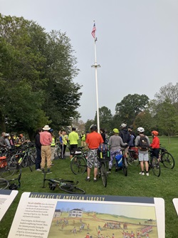 People and bikes gathered on Lexington Green in front of a statue and flagpole. Signage describing the site is in the foreground.